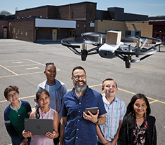 Photo of Antonio Saccucci, Ontario Certified Teacher, outside in the school playground with five students. All are smiling. There is a drone hovering over their heads.